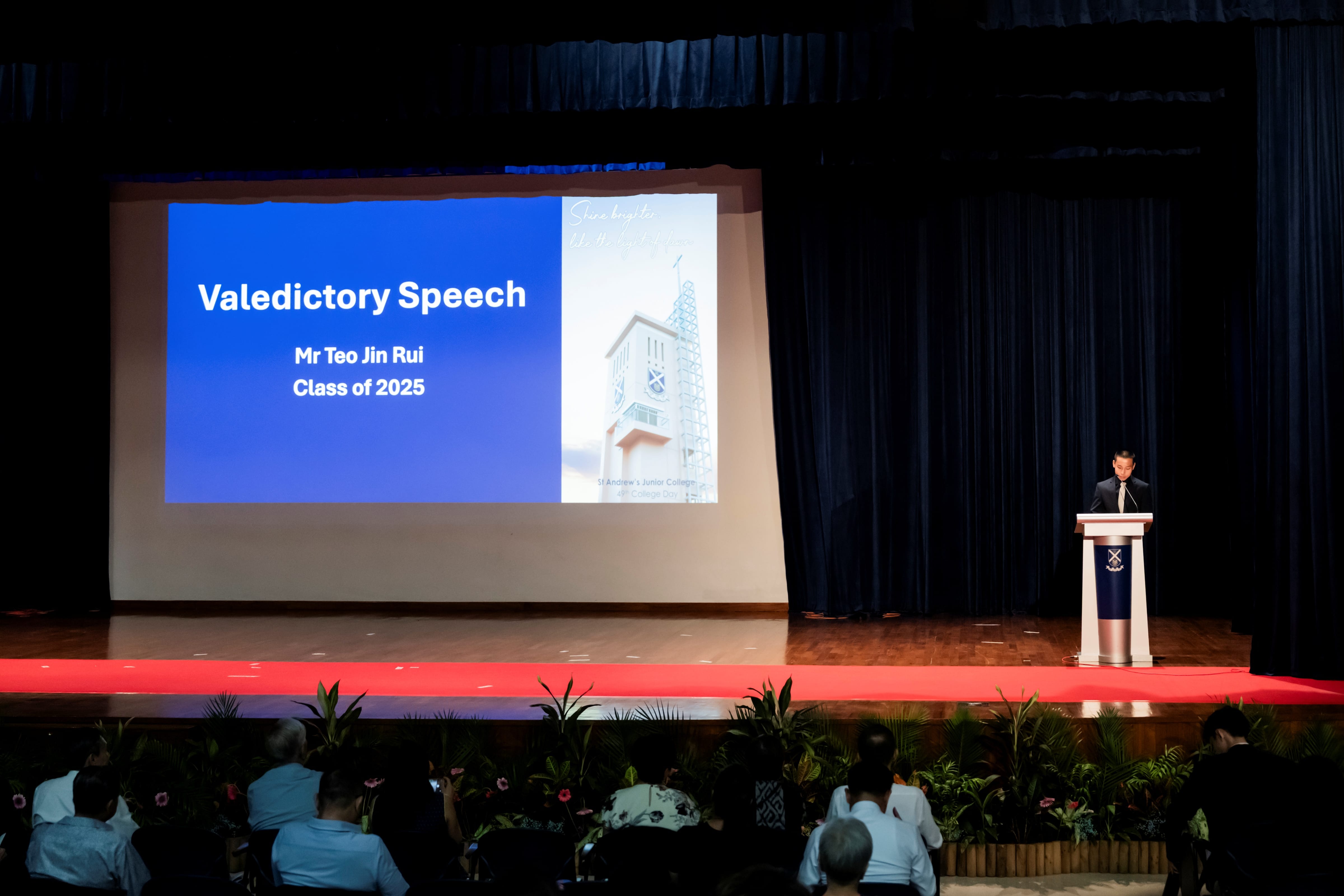Valedictorian of the Class of 2025, Teo Jin Rui, stands at a podium on stage beside a large projection screen displaying "Valedictory Speech: Mr Teo Jin Rui, Class of 2025."